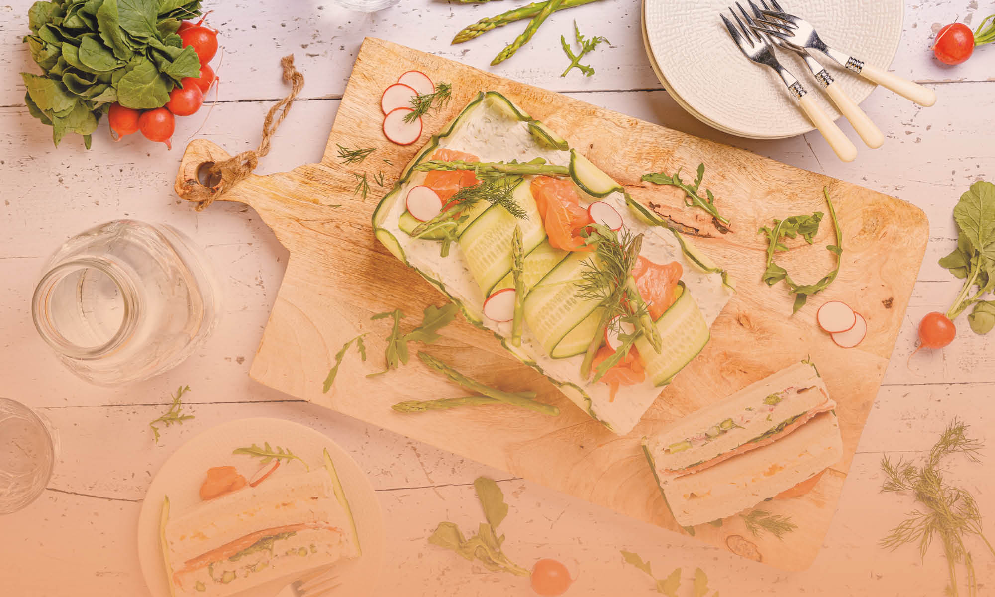 Een houten snijplank met een sandwich versierd met dunne plakjes komkommer, asperges, radijs, dille en gerookte zalm. Naast de plank liggen verse groenten zoals radijsjes en rucola, een glazen karaf met water, een bord met een sandwich en een stapel witte borden met drie vorken erop, alles op een lichte houten tafel.